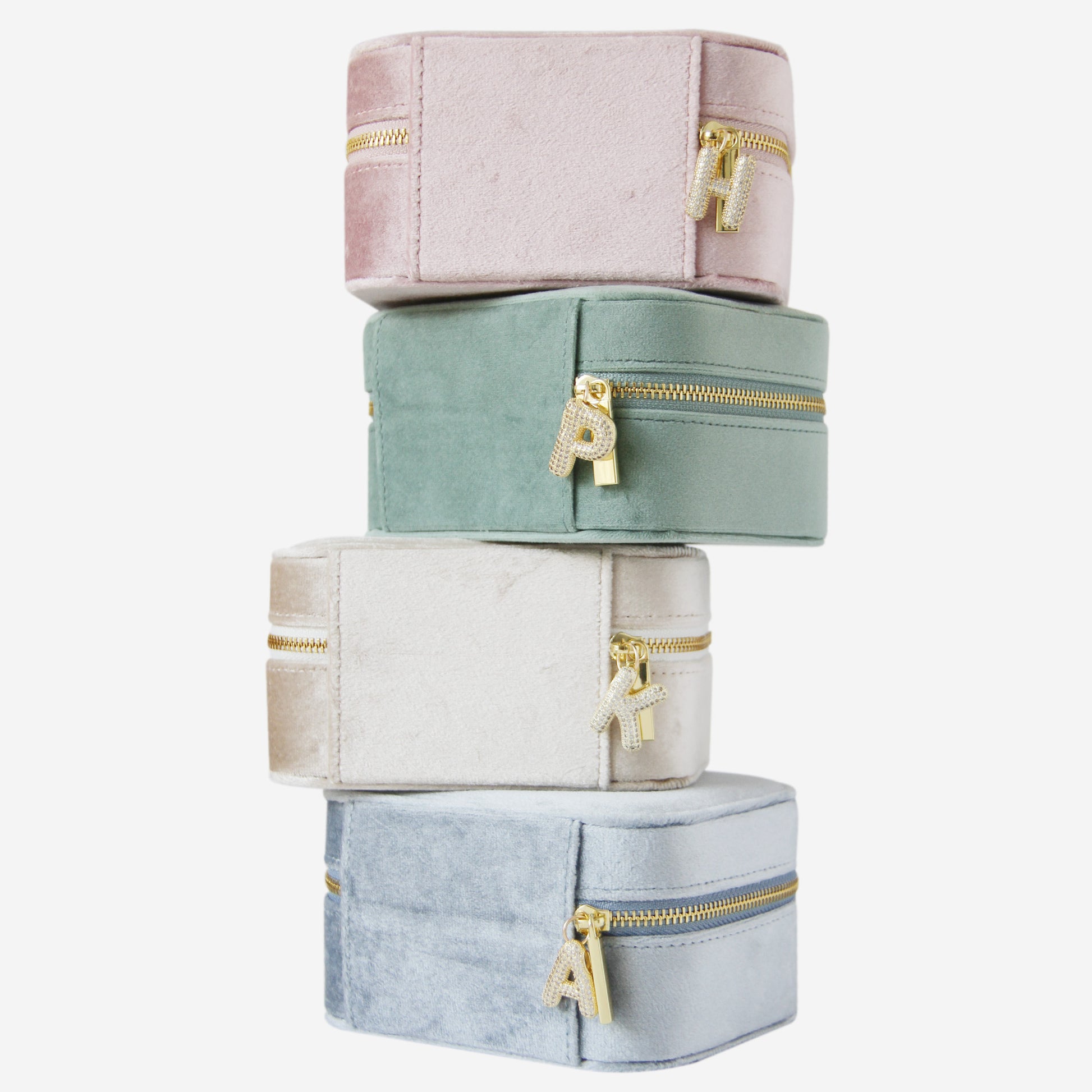 Stack of four pastel-colored jewelry boxes with gold zippers and gold and crystal letter charms on a white background